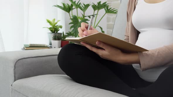 Mid section of a pregnant woman sitting cross-legged on a couch writing in notebook