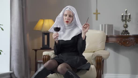Wide Shot Portrait of Slim Gorgeous Caucasian Woman in Nun Costume Sitting Indoors with Wine Glass alt