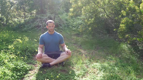 A man sitting in a meditation pose in a green sunny forest meadow practicing deep breathing exercise alt