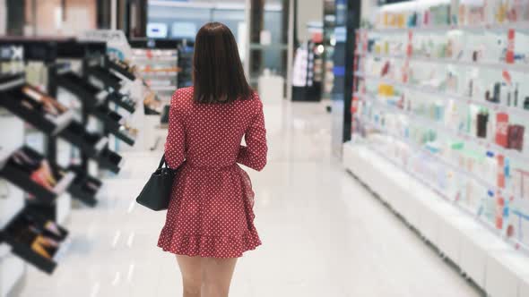 Brunette Woman in Red Dress Walks Along Shelves in Cosmetic Boutique alt