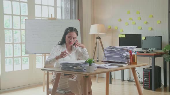 Asian Woman Talking On Phone And Taking Note While Working Hard With Documents At Office alt