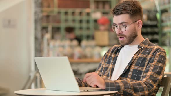 Young Man Celebrating Success on Laptop in Cafe  alt