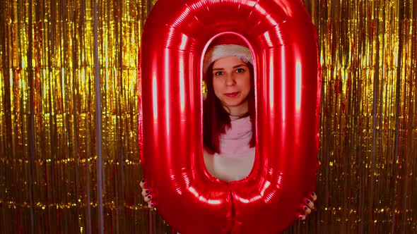 Woman in Santa Hat Holds Red Figure Zero on Background of Shimmering Golden Tinsel alt