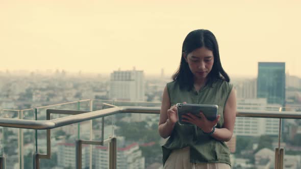 Beautiful woman using tablet chatting with friends while standing beside a balcony on a rooftop.