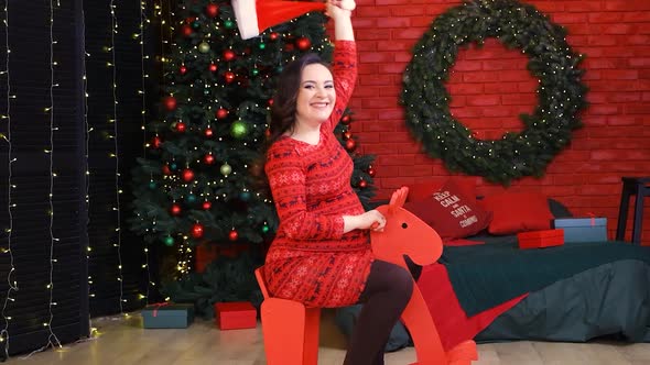 A Happy Pregnant Woman Rides a Red Wooden Toy Horse and Has Fun Preparing for Christmas alt