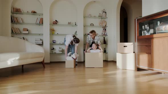 Excited Happy Young Parents Run Push Cardboard Boxes with Little Cute Girl Daughters Ride Insideand alt