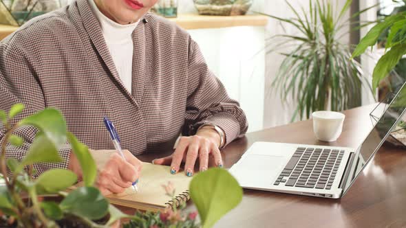 Senior Woman in Glasses Sitting with Laptop and Writing in Notebook. alt