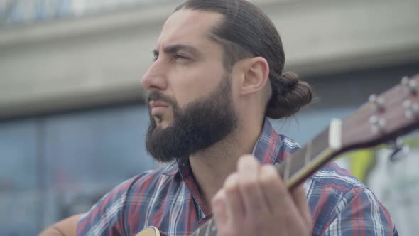 Close-up Portrait of Engrossed Caucasian Musician Playing Guitar Outdoors. Handsome Absorbed Man alt