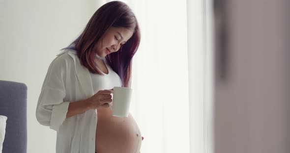 Asian pregnant woman drinking water while standing beside windows and touching her belly.