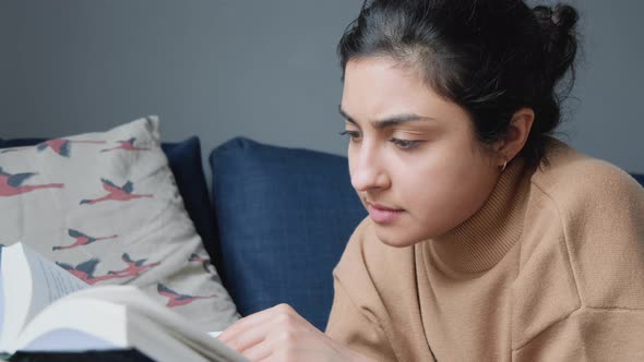 Close Up of a Young Indian Woman Sitting Next to a Sofa Thoughtfully Reading a Book alt