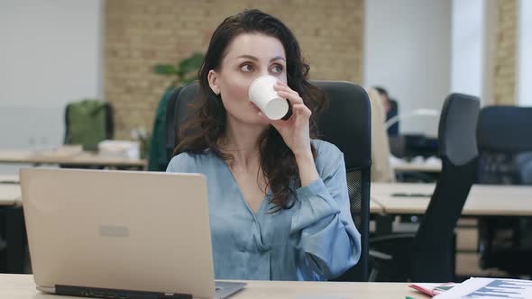 Charming Successful Businesswoman Typing on Laptop Keyboard and Drinking Coffee in Office. Portrait alt
