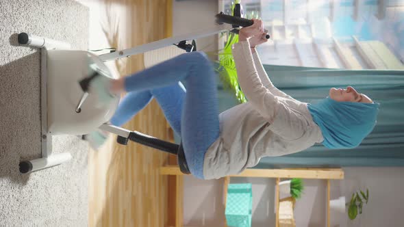 Muslim Woman in a National Headscarf is Engaged on an Exercise Bike in the Living Roomvertical Video alt
