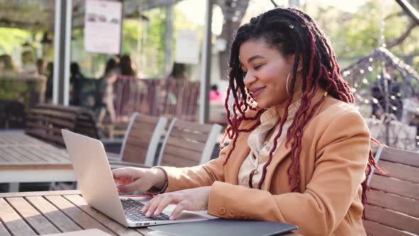 Young African American Woman Sitting in Outdoor Cafe and Using Laptop