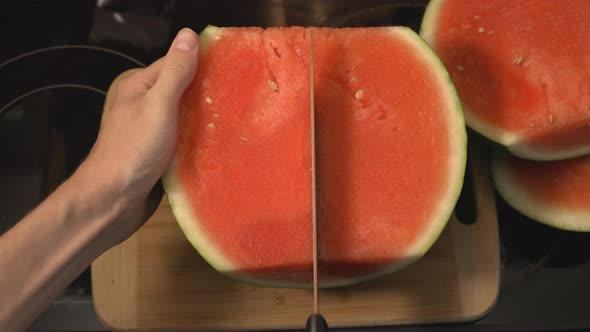 Hand Firmly Holding A Half-Sliced Watermelon Side Down Slicing In Half Lengthwise With A Sharp Knife alt