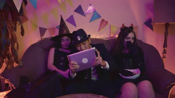 A Guy in a Creepy Costume is Actively Playing on a Tablet and the Girls are Sitting and Bored at a alt