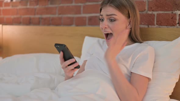 Cheerful Young Woman Celebrating Success on Smartphone in Bed  alt