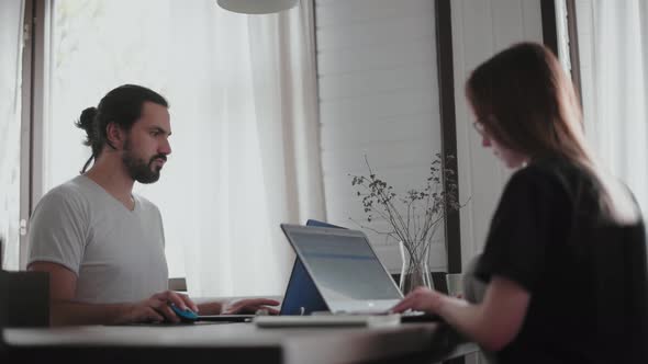 A young man and a young girl are working in front of laptops in their bright apartment. alt
