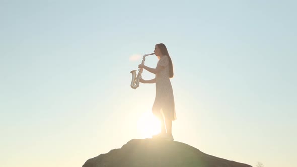 Beautiful Woman in Light Wind Blown Dress Plays Saxophone Against Backdrop of Sunset alt