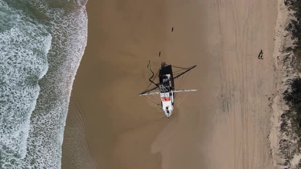 High drone view looking down at a fishing trawler recently washed up on a deserted beach with waves alt