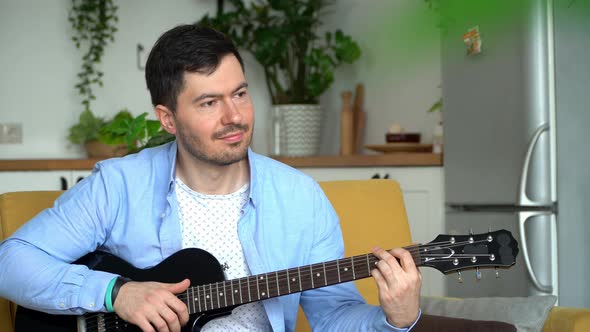 Portrait of Young Smiling Man Sits on Couch at Home and Plays Electric Guitar
