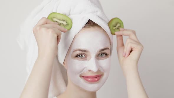 Young Woman with White Mud Mask on Face on White Background Posing with Kiwi Slices and Smiling alt