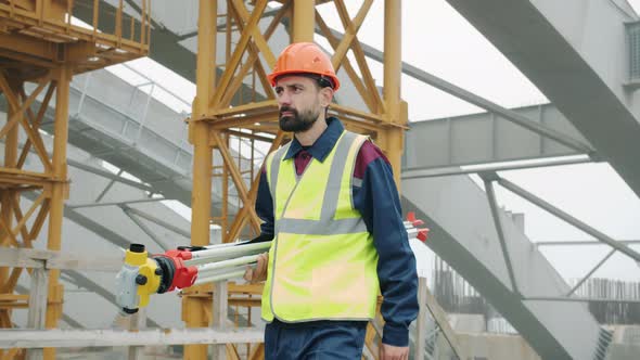 Dolly Shot of Serious Man Surveyor Carrying Theodolite Walking in Building Site Alone alt