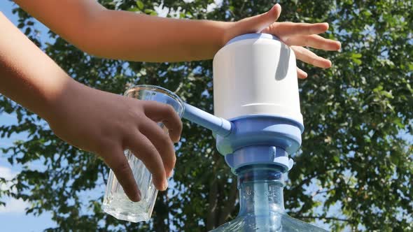 Hands of a Boy Teenager Pump Drinking Water Into a Glass Cup with a Mechanical Plastic Pump on a alt
