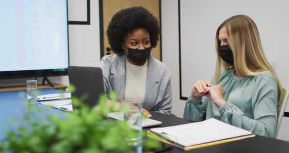Two diverse businesswomen wearing face masks working together using laptop at desk in office alt