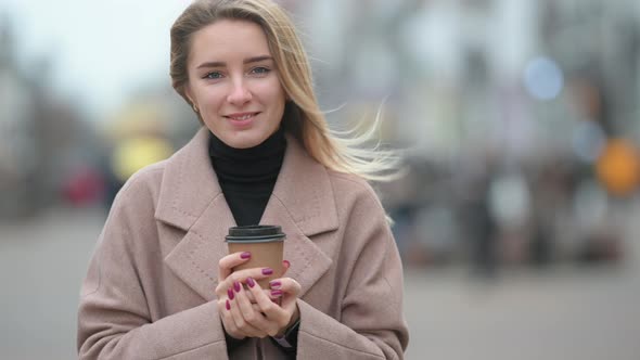 Portrait of a young girl in a coat standing on street and holding cup of hot drink.