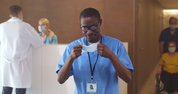 Afroamerican Male Nurse Putting on Safety Mask Looking at Camera Standing in Hospital Waiting Room alt