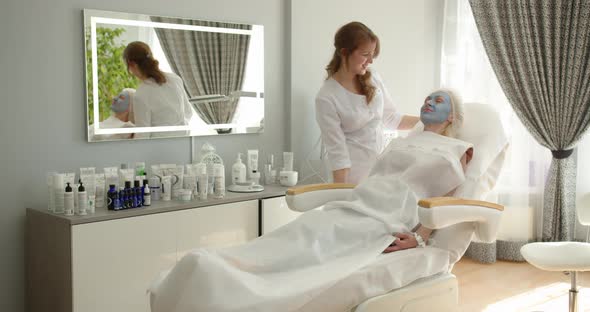 Cosmetology Modern Salon. A Beautician Lowers A Beauty Chair With A Girl In A Blue Clay Mask alt