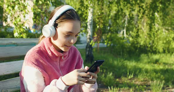 Ten Years Old Girl with Smartphone and in Headphones  She Chatting with Friends Outdoor alt