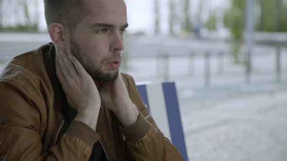 Upset Young Man Sitting on Wooden Bench and Rubbing Head alt