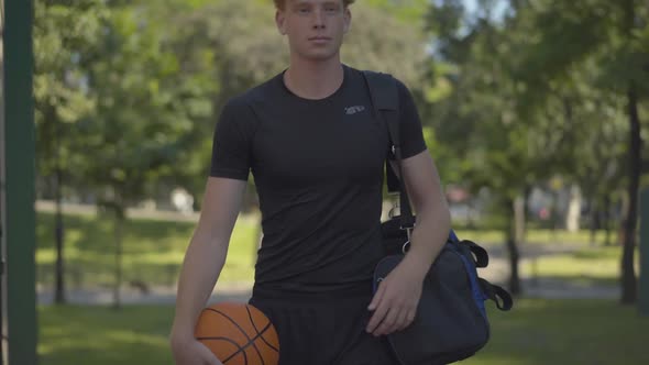 Portrait of Smiling Confident Sportsman Entering Outdoor Court with Sports Bag and Ball. Positive alt
