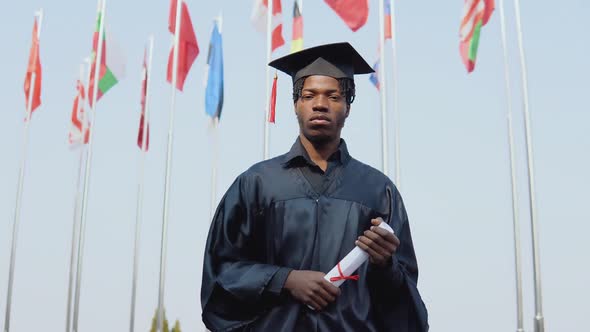 Young African American Male Graduate Standing in Front of the Camera with a Diploma and Books in Her alt
