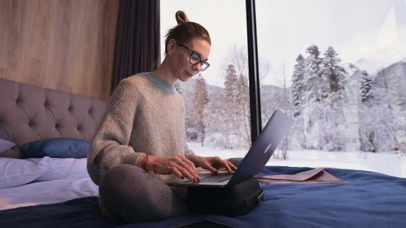 Portrait of Attractive Freelancer Woman with Glasses and a Sweater with Stockings Sitting on a Bed alt