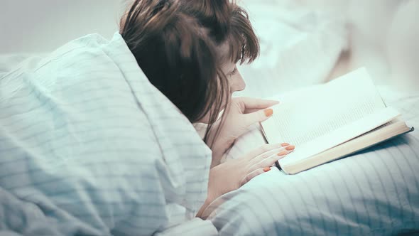 Pretty Brunette Woman Lies In Bed And Attentively Reads A Book View From Above