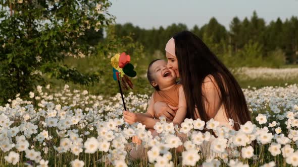 Slow Motion of Young Attractive Mother with Son Playing Windmill Sitting in Flower Field at Sunset.