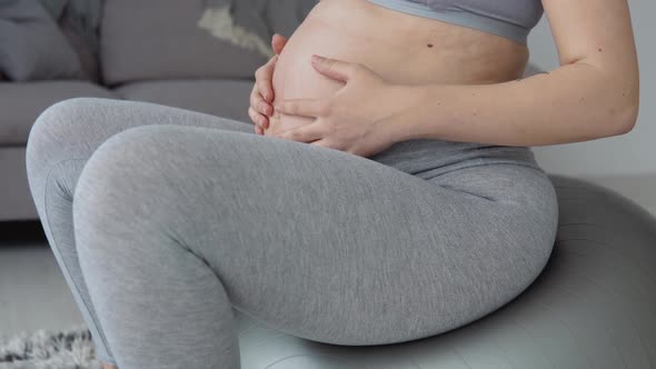 A Pregnant Woman in Sportswear Sits on a Fitball and Holds Her Hands on Her Stomach alt