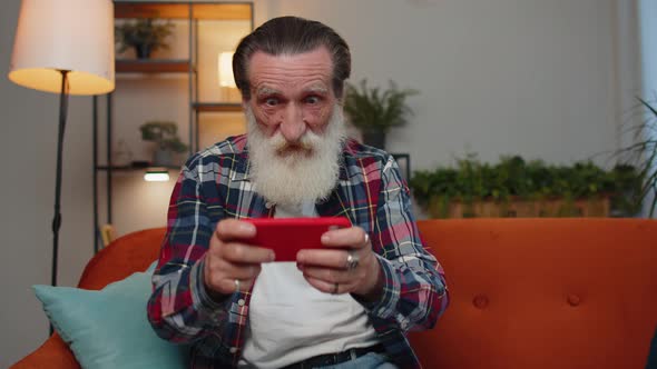 Worried Funny Senior Old Grandfather Man Playing Racing Online Video Games on Smartphone at Home alt
