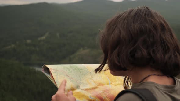 A young girl on a trip stands with a paper map against the background of mountains and a lake.