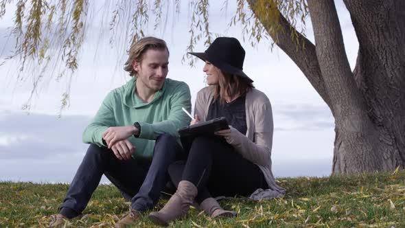 Young couple sitting under tree looking at tablet and talking alt