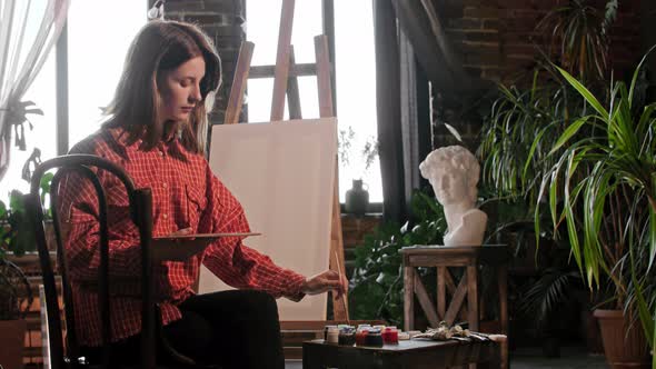 A Young Pretty Woman Artist Mixing Colors on the Palette Sitting in Front of an Easel alt