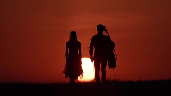 Silhouette of Woman Dancer and Cellist Walking Opposite Huge Sun
