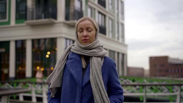the Blonde Is Dressed in a Blue Coat and a Gray Scarf on Her Head Walking Down a City Street. She alt