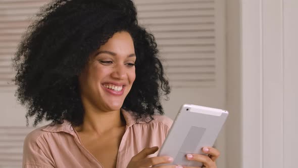 Cute African American Woman Looks Information on Digital Tablet and Happily Celebrates Victory alt