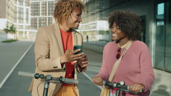 Afro Man and Woman with E-Scooters Using Phone and Talking on Street alt