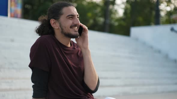 Portrait of Young Attractive Arab Man Sitting on the Grass and Talking on the Phone alt