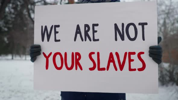 A Protester is Standing in a Park with a Sign We Are Not Your Slaves. Picket. alt