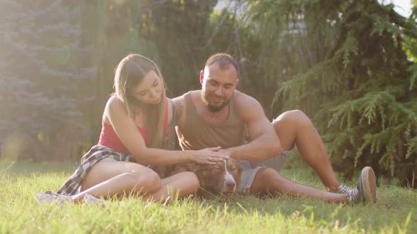 Young Couple Sitting in Park on Grass and Petting Their Dog on Sunny Summer Day